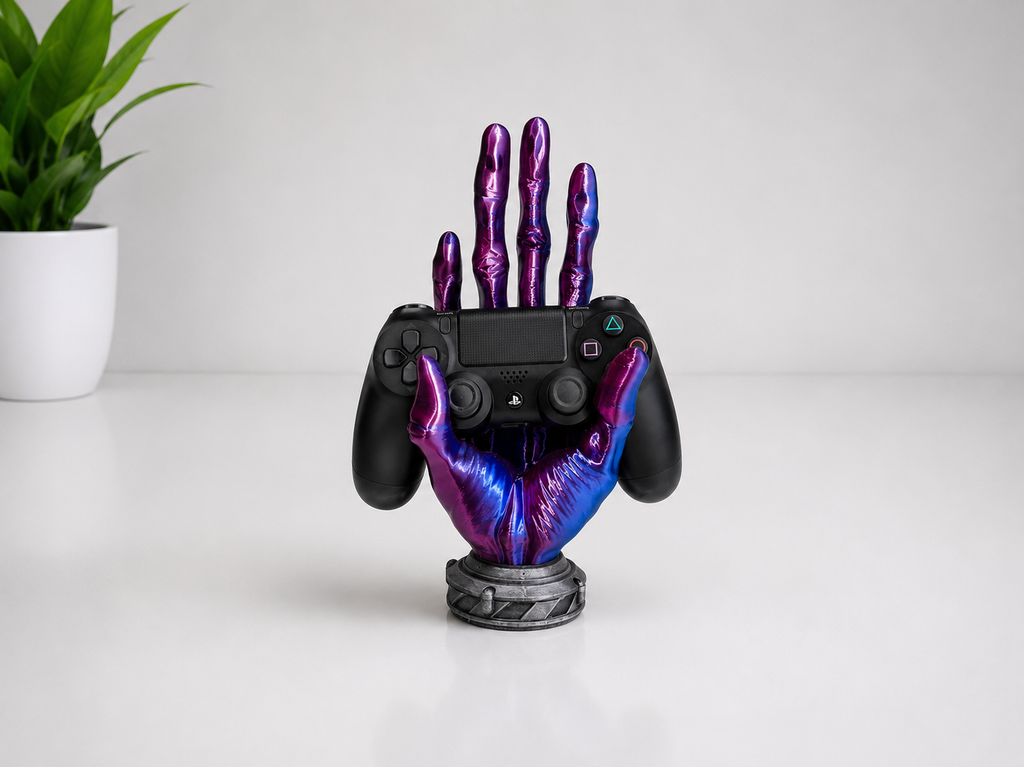 A black PlayStation 5 controller held upright by a decorative metallic hand-shaped stand with a purple and blue iridescent finish, set on a white surface with a small potted plant in the background.