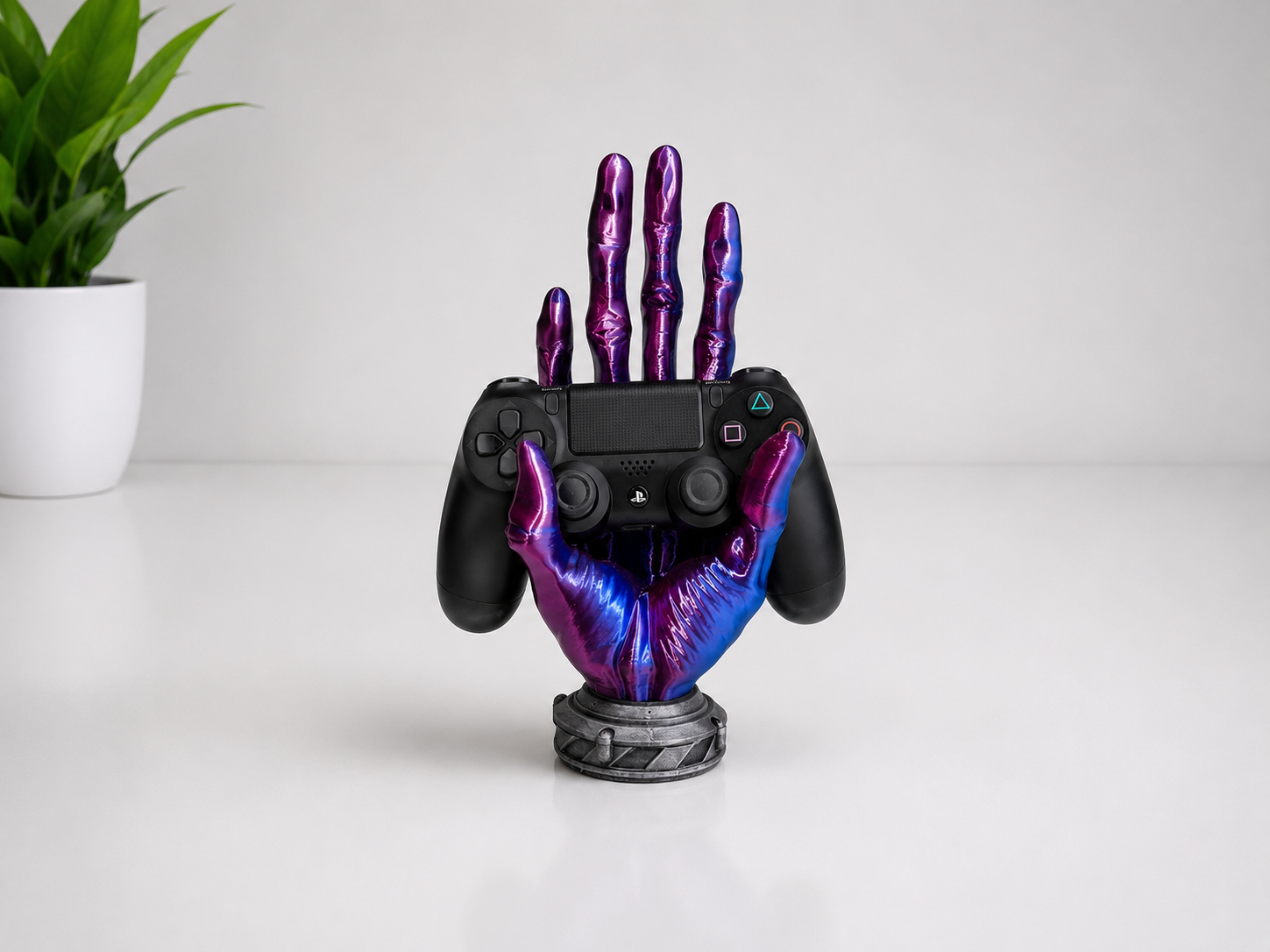 A black PlayStation 5 controller held upright by a decorative metallic hand-shaped stand with a purple and blue iridescent finish, set on a white surface with a small potted plant in the background.