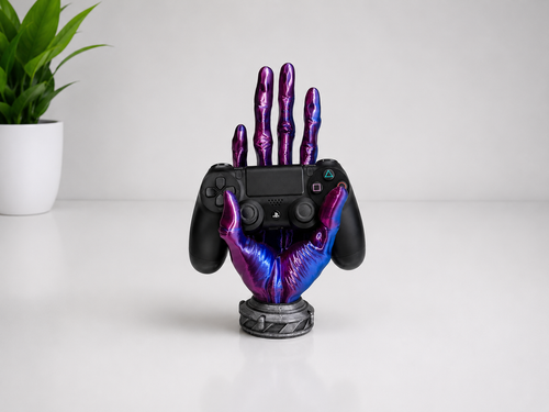 A black PlayStation 5 controller held upright by a decorative metallic hand-shaped stand with a purple and blue iridescent finish, set on a white surface with a small potted plant in the background.