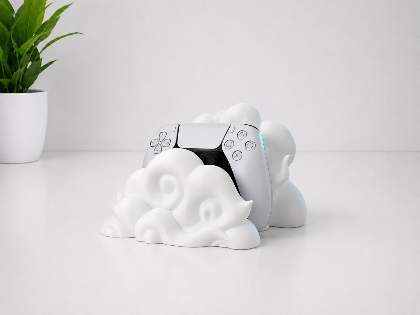 A white cloud-shaped stand holding a white PlayStation 5 controller, set on a clean white surface with a small green potted plant in the background.