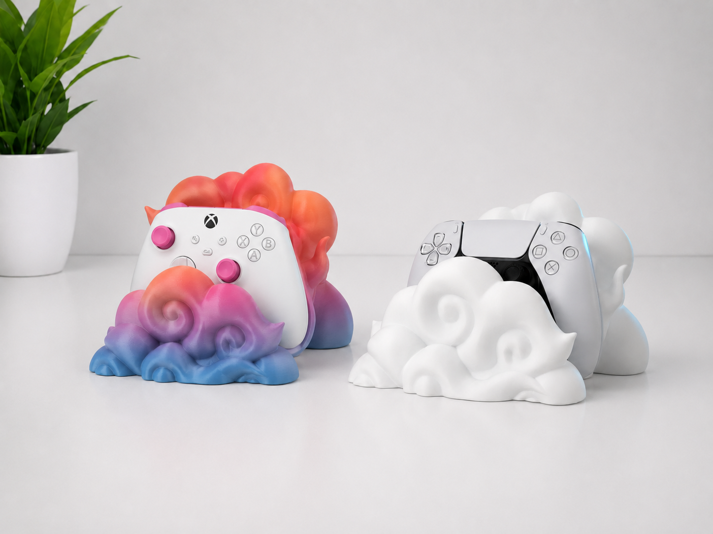Two game controllers resting on decorative cloud-shaped stands, one with a colorful gradient cloud base and one in white, set against a clean white background with a small potted plant in the corner.
