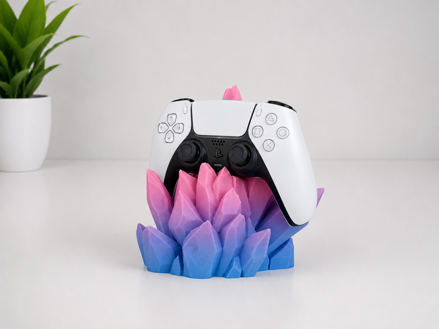 A white and black PlayStation 5 DualSense controller displayed on a colorful 3D-printed stand shaped like a lotus flower, set on a white surface with a small potted plant in the background.