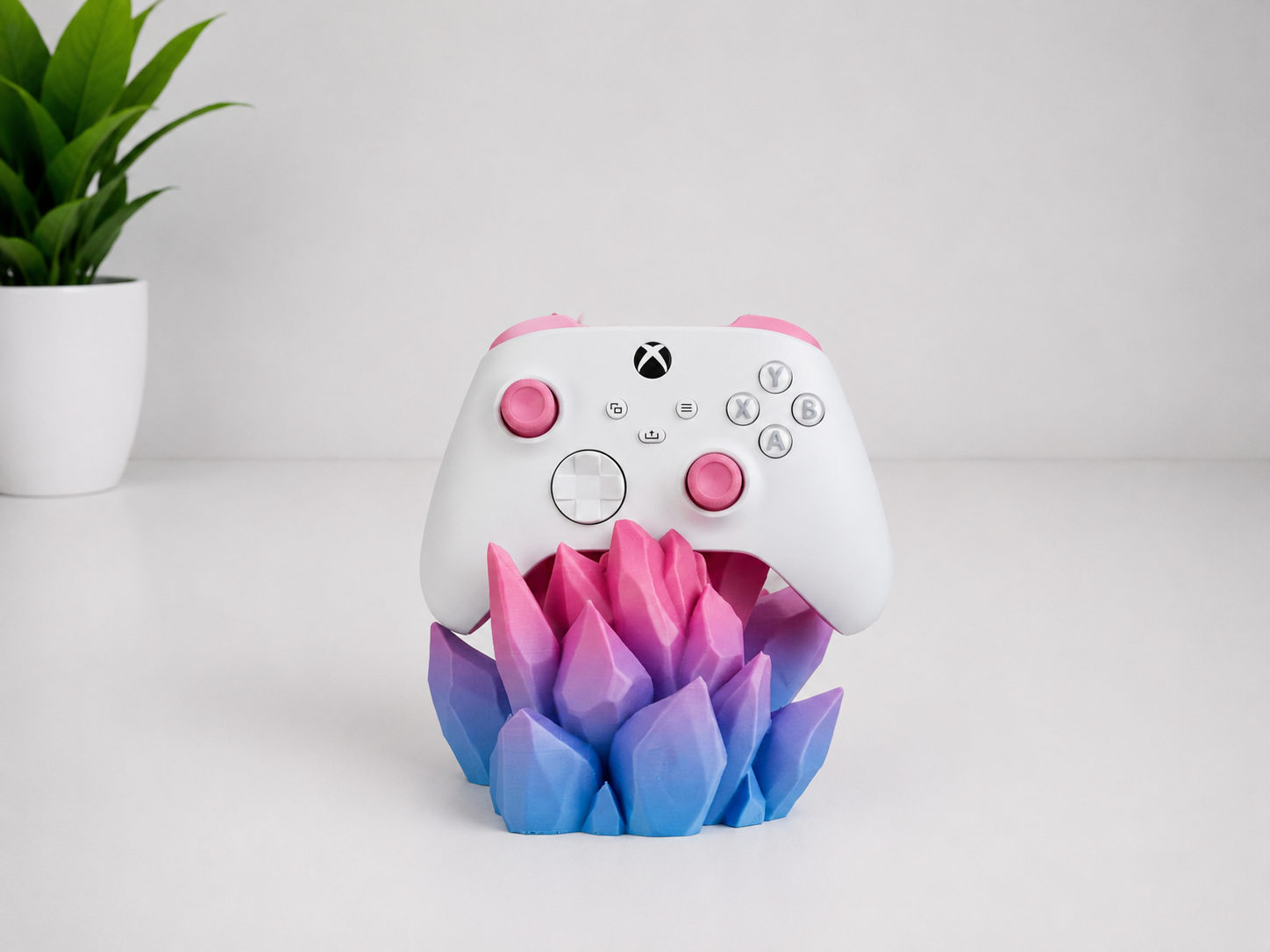 A white Xbox controller with pink accents displayed on a colorful, lotus-shaped stand that transitions from pink to purple to blue, set against a clean white background with a small green plant in the corner.