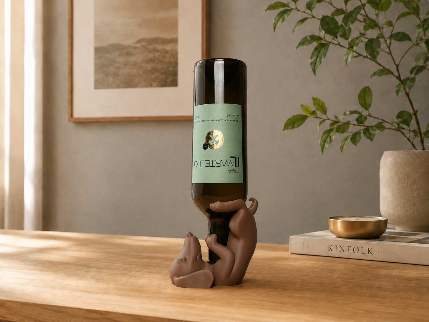 A dark glass wine bottle with a light green label is held upside down by a small brown dog figurine on a wooden table. The scene is styled in a warm, modern interior with a potted plant, a book, and a small bowl in the background.