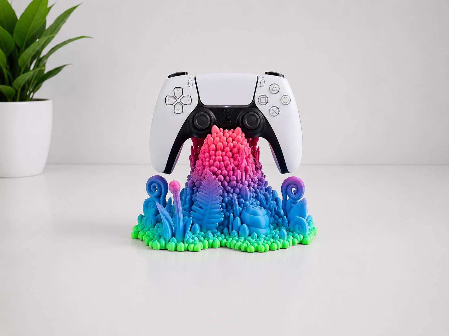 A white PlayStation 5 DualSense controller sits centered on a colorful 3D-printed stand shaped like a mushroom cap, with a small green potted plant in the background on a white surface.