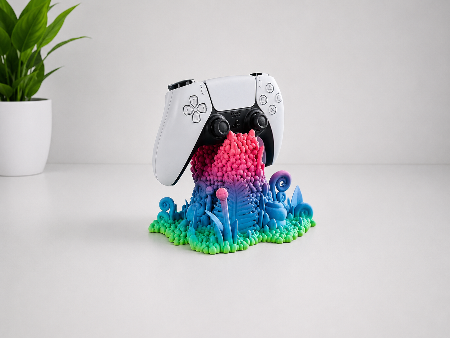 A white and black PlayStation 5 controller resting on a colorful, sculptural 3D-printed stand with a vibrant blue, pink, and green gradient. The stand is placed on a clean white surface with a small potted plant in the background.