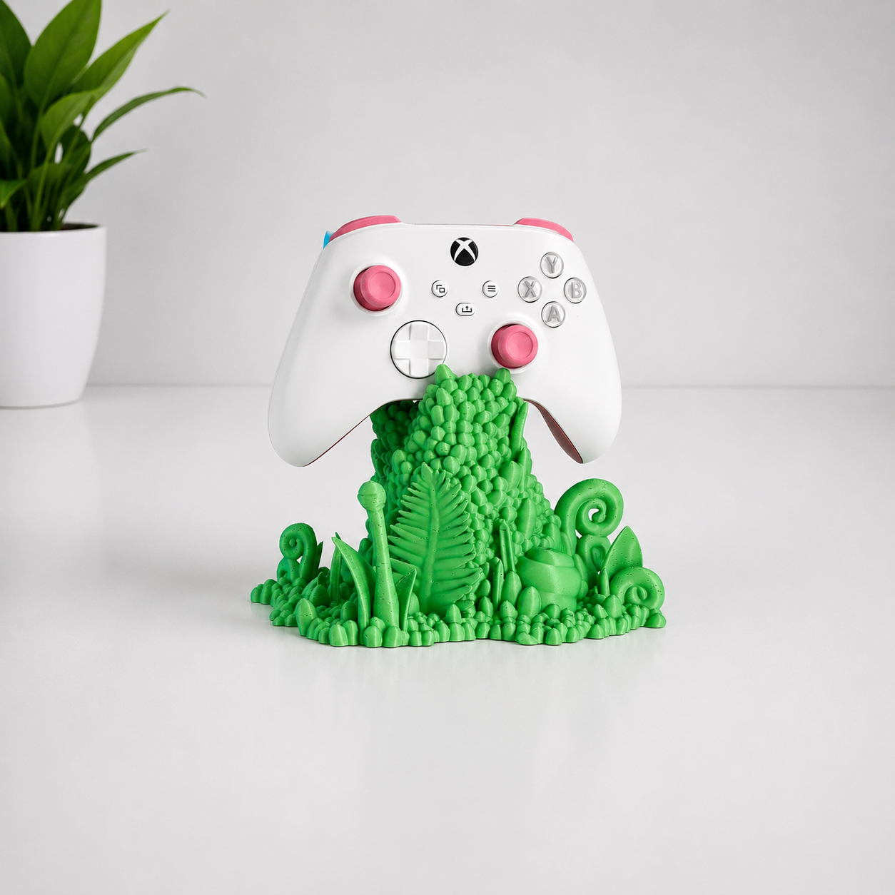 A white Xbox controller with pink joysticks and accents sits on top of a green jungle-themed silicone stand featuring a fern, turtle, and leaf motifs, set on a white surface with a blurred potted plant in the background.