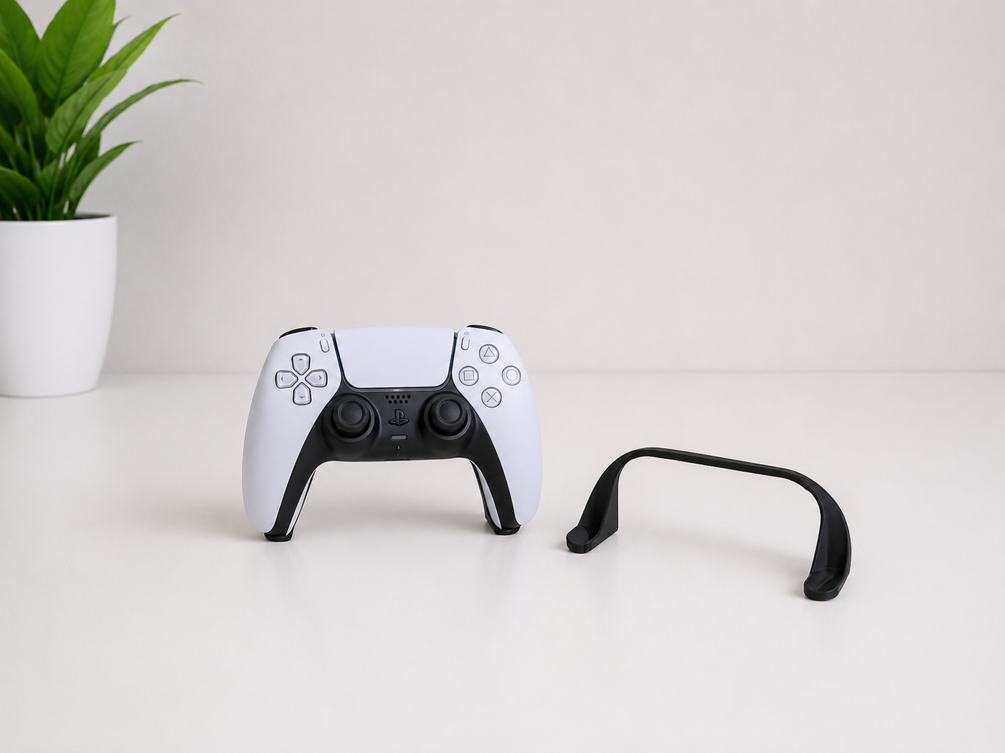 A white and black PlayStation 5 DualSense wireless controller standing upright on a white surface, with a black curved accessory placed to its right. A small green potted plant is visible in the background against a clean white wall.