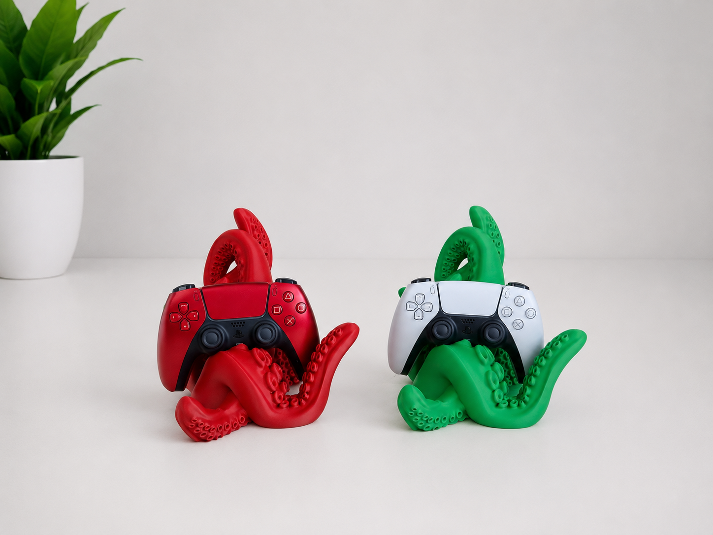 Two octopus-shaped game controller stands in red and green, each holding a PlayStation-style controller, on a white surface with a small potted plant in the background.