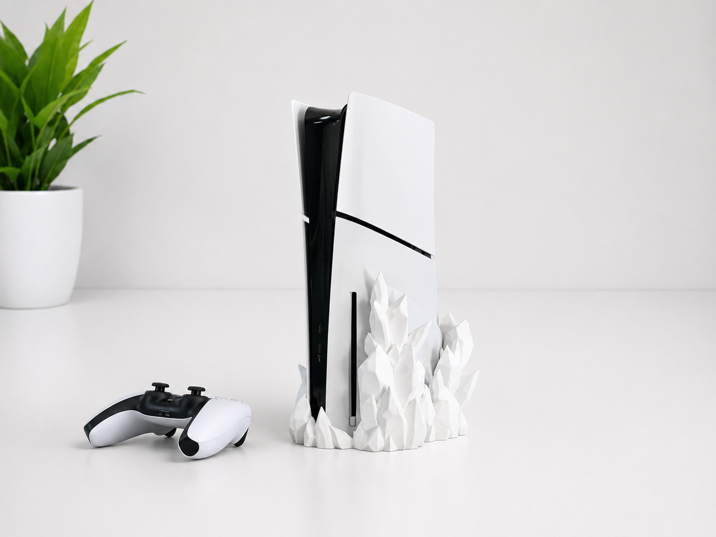 A white PlayStation 5 console with a black front panel stands on a white sculptural base, with a matching white and black controller placed to the left on a clean white surface. A small green potted plant is visible in the background.