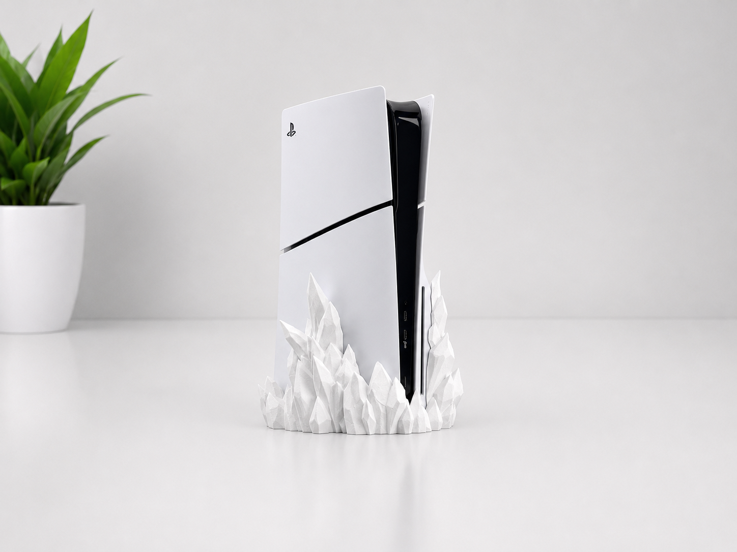 A white PlayStation 5 console standing upright on a sculpted white mountain-like base, set on a light gray tabletop with a small green potted plant in the background.