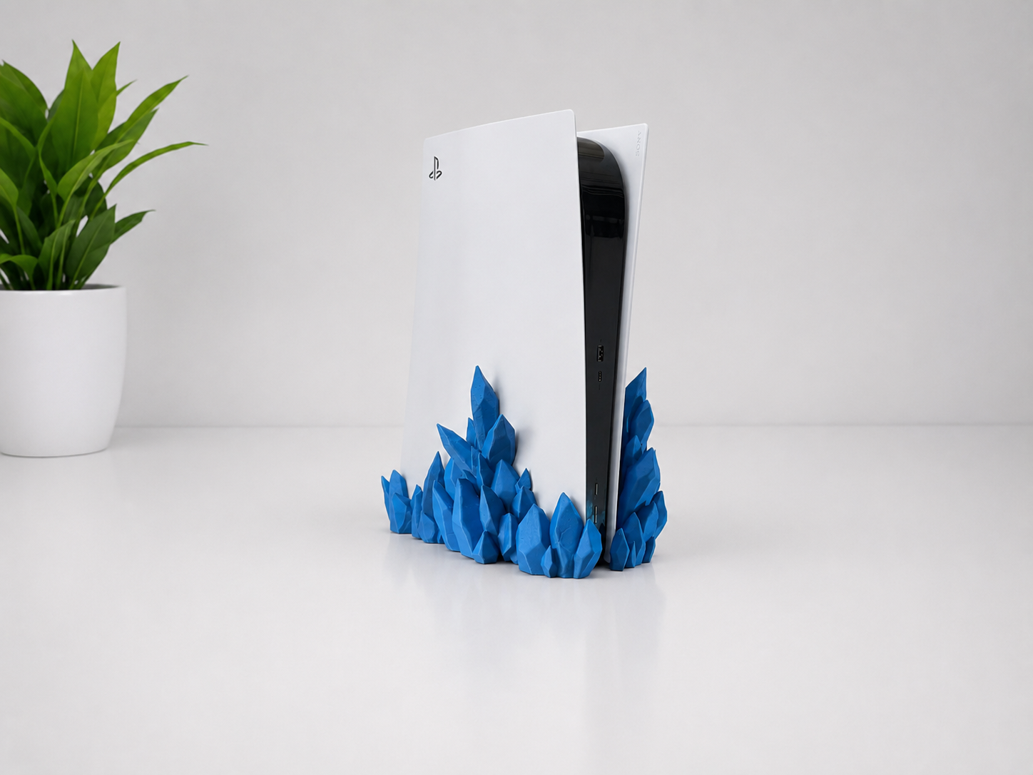 A PlayStation 5 console in white and black standing upright on a blue faceted, crystal-like base, photographed on a clean white surface with a small green plant in a white pot at the left edge.
