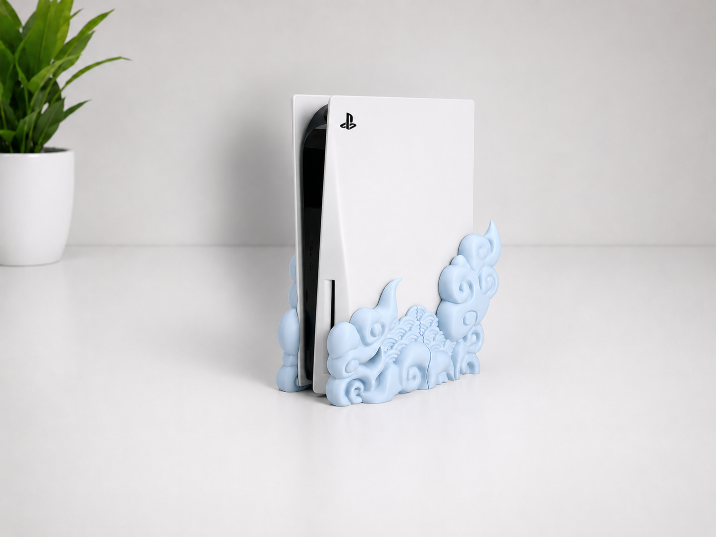 A white PlayStation 5 console standing upright in a decorative light blue cloud-shaped holder on a white surface, with a small potted green plant in the background.