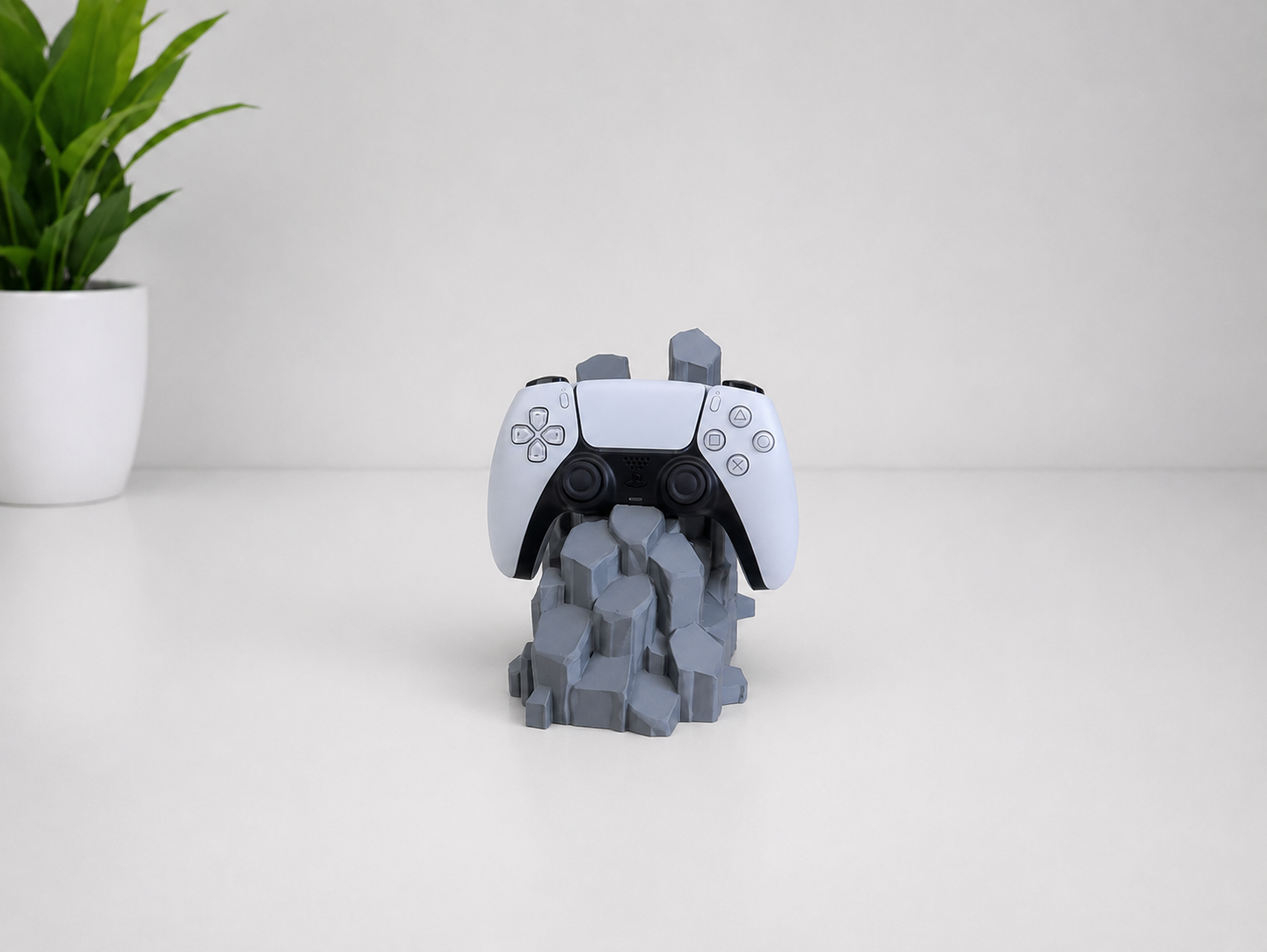 A white and black PlayStation 5 DualSense controller displayed upright on a gray blocky, rock-like stand against a clean white background, with a small green potted plant in the upper left corner.