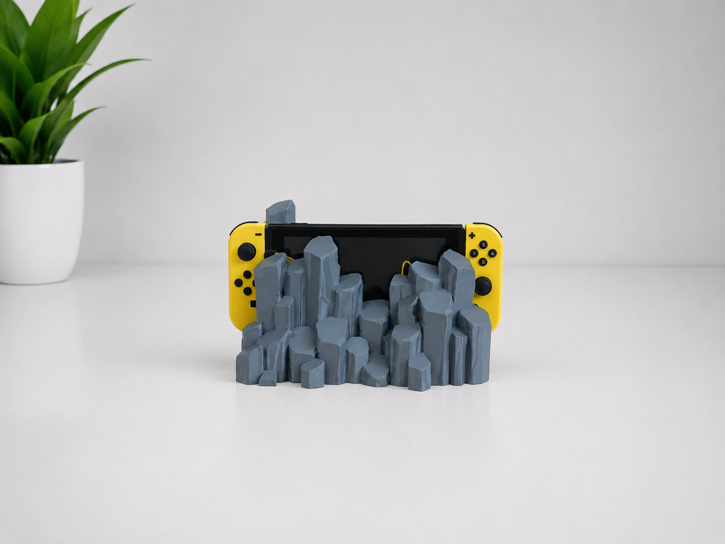 A gray 3D-printed quartz-shaped stand holding a yellow Nintendo Switch, set on a white surface against a light gray background with a small green plant in the corner.