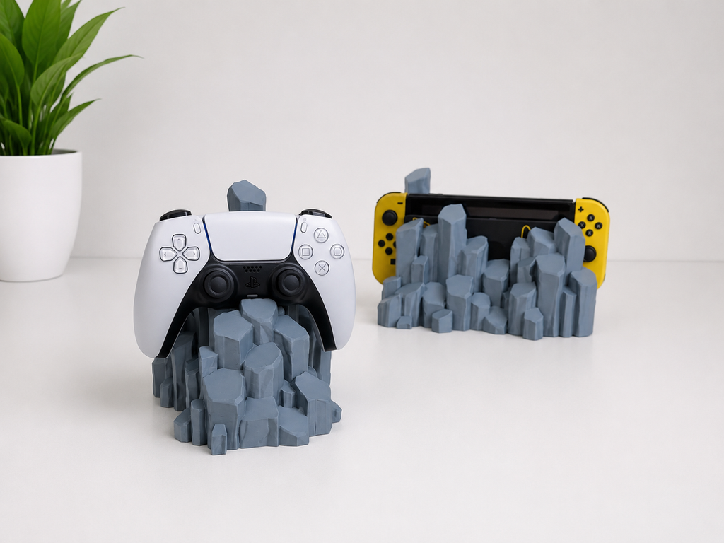 A gray 3D-printed quartz-shaped game controller stand holding a white PlayStation 5 controller, with a second stand holding a Nintendo Switch console in yellow Joy-Con controllers, shown on a white surface with a small potted plant in the background.