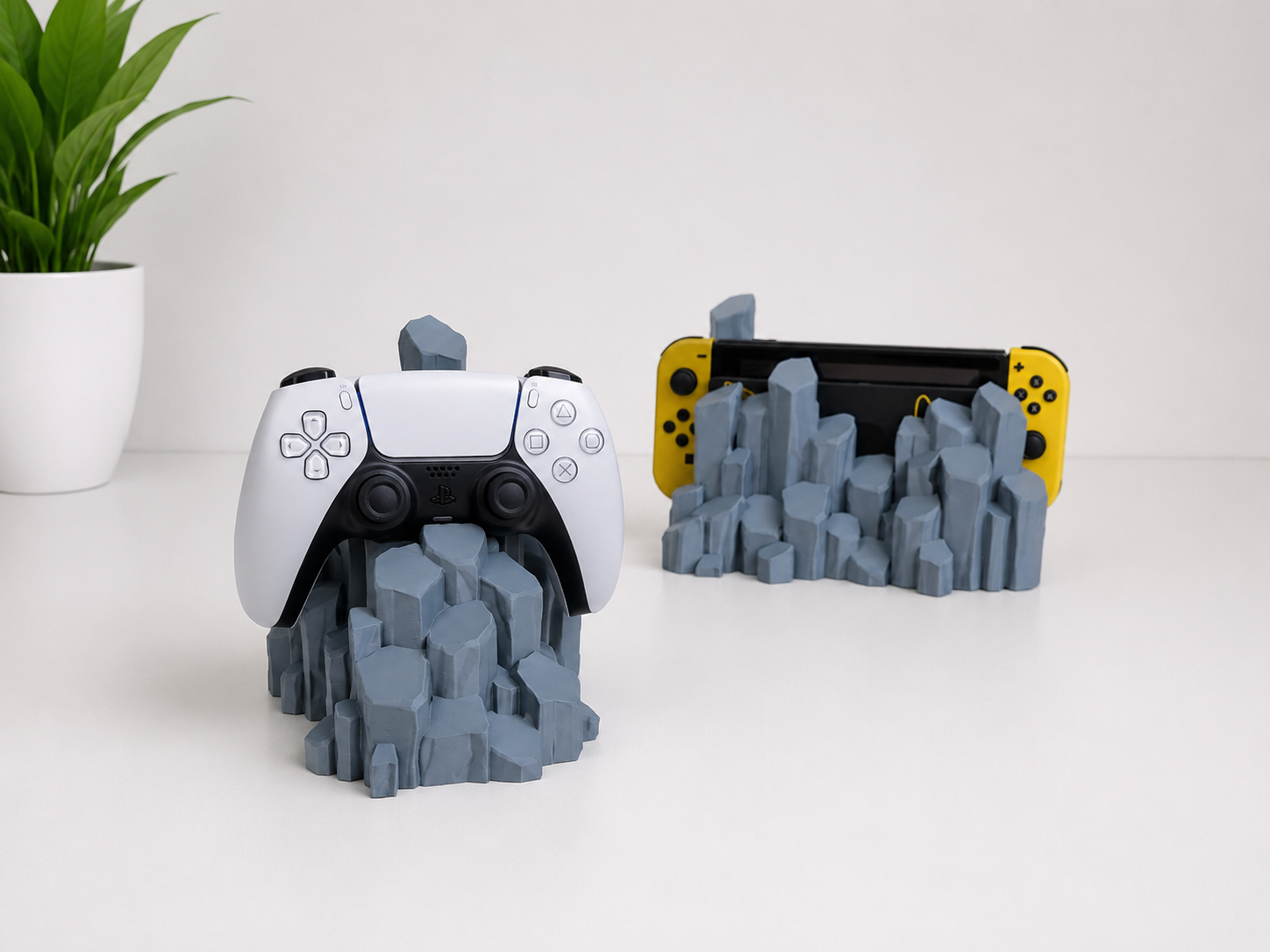 A gray 3D-printed quartz-shaped game controller stand holding a white PlayStation 5 controller, with a second stand holding a Nintendo Switch console in yellow Joy-Con controllers, shown on a white surface with a small potted plant in the background.