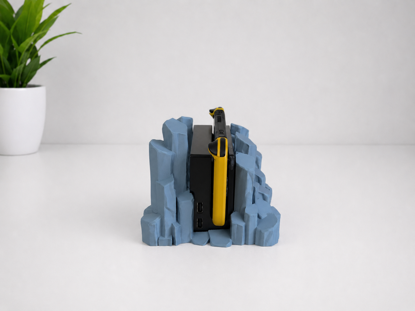 Side view of a gray 3D-printed quartz-shaped stand holding a yellow Nintendo Switch, set on a white surface against a light gray background with a small green plant in the corner.