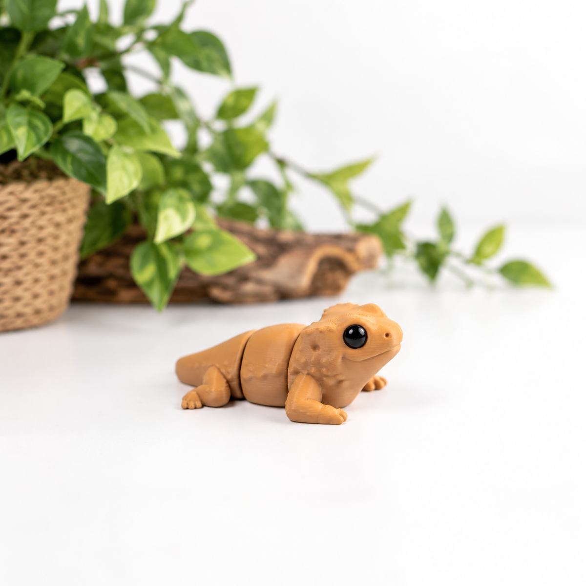 A small tan lizard figurine with a glossy black eye, positioned on a white surface with a blurred green potted plant in the background.