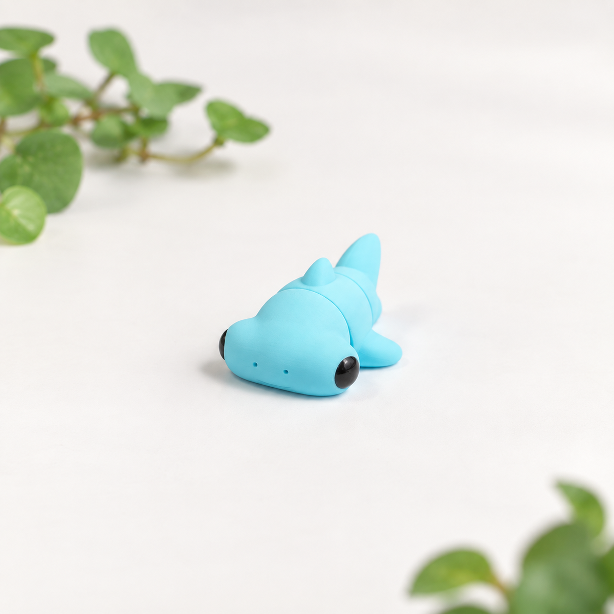 A small light blue shark-shaped object with black eyes, photographed on a clean white surface with soft green plant leaves in the background.