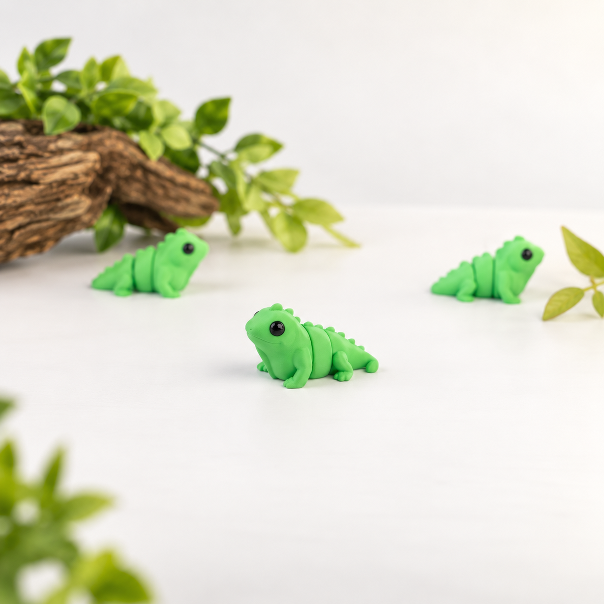 Three small iguanas figurines with black eyes, arranged on a white surface with decorative green leaves and a piece of driftwood in the background.