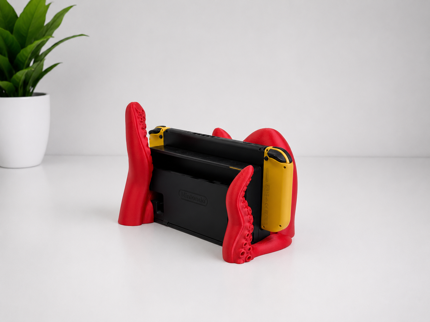 A red and black 3D-printed octopus tentacle game controller stand holding two yellow and black Nintendo Switch controllers, set on a white surface with a potted plant in the background.