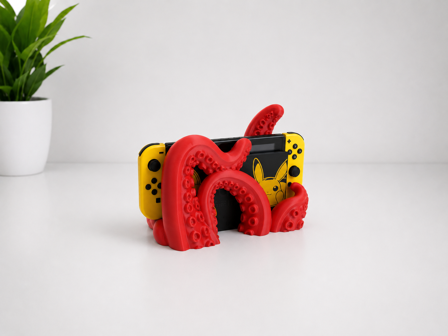 A red switch stand designed with a tentacle pattern, holding a black Nintendo Switch with yellow Joy-Con controllers. The setup is on a clean white surface with a small green potted plant in the background.